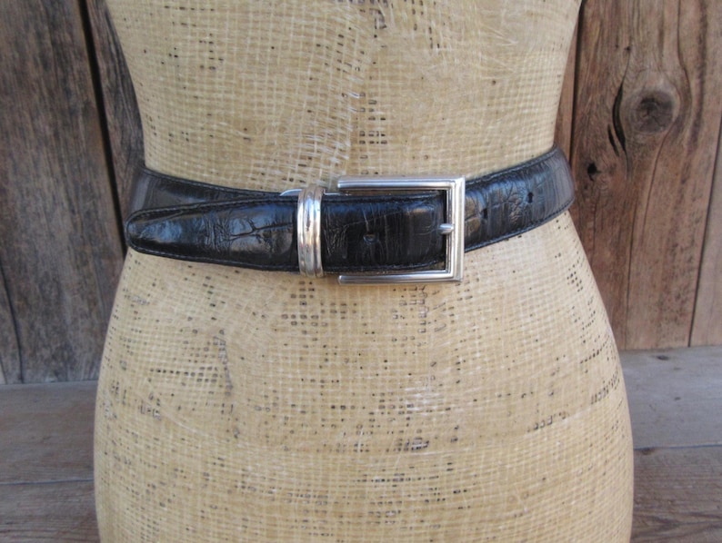 May include: A black leather belt with a silver rectangular buckle. The belt has a textured, embossed pattern. The buckle is a simple, classic design. The belt is displayed on a mannequin.