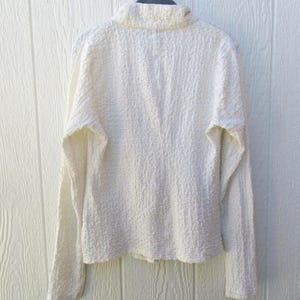 May include: A long-sleeved, off-white shirt featuring a high collar and a distinctively textured, crinkled fabric surface. The garment is displayed on a dark hanger, showcasing a solid, neutral color and a structured, fitted silhouette.