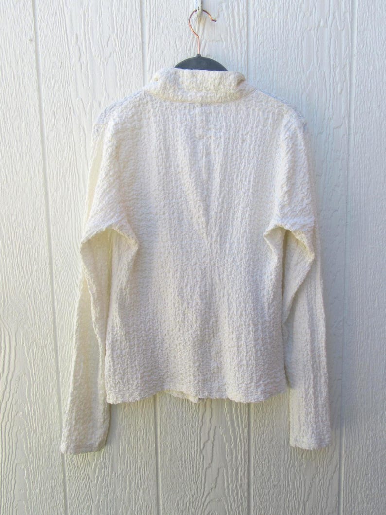 May include: A long-sleeved, off-white shirt featuring a high collar and a distinctively textured, crinkled fabric surface. The garment is displayed on a dark hanger, showcasing a solid, neutral color and a structured, fitted silhouette.