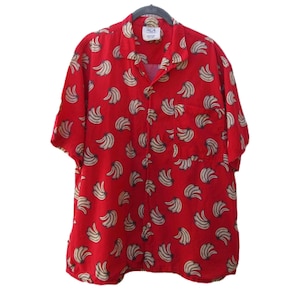 May include: Red short-sleeved button-down shirt with a repeating banana pattern. The shirt has a collared neckline, a chest pocket, and is hanging on a black hanger. The bananas are cream-colored with brown accents.
