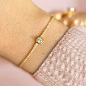 May include: A fine gold bracelet with a woven chain. It has a single, round, clear gemstone set in a gold bezel. The bracelet is shown on a wrist, with a pink garment visible in the background.