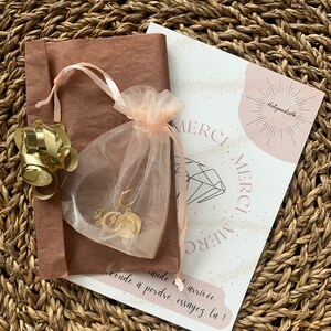 K&ouml;nnte beinhalten: Ein goldfarbenes Schmuckset, pr&auml;sentiert in einem durchsichtigen rosa Beutel und einer braunen Papierverpackung, gebunden mit einem rosa Band. Eine Karte mit dem Wort "MERCI" ist ebenfalls enthalten, alles auf einem gewebten Hintergrund.