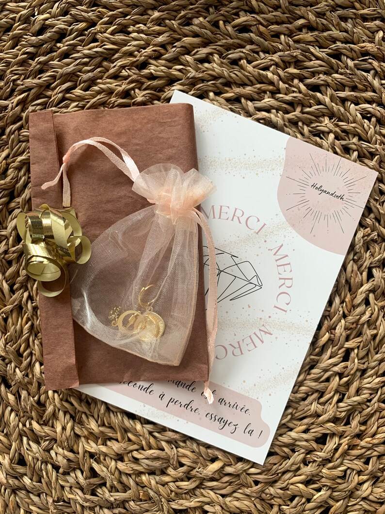 K&ouml;nnte beinhalten: Ein goldfarbenes Schmuckset, pr&auml;sentiert in einem durchsichtigen rosa Beutel und einer braunen Papierverpackung, gebunden mit einem rosa Band. Eine Karte mit dem Wort "MERCI" ist ebenfalls enthalten, alles auf einem gewebten Hintergrund.