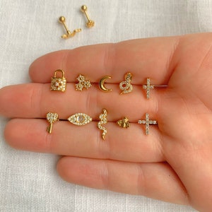 May include: A collection of gold-colored stud earrings in various designs, including a lock, flower, crescent moon, snake, cross, key, eye, bee, and a pair of screw-back earrings. Some earrings are adorned with small, clear gemstones.