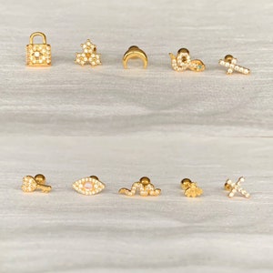 K&ouml;nnte beinhalten: Eine Sammlung goldfarbener Ohrringe mit verschiedenen Designs, darunter ein Schloss, Blumen, ein Halbmond, eine Schlange, ein Kreuz, ein Schl&uuml;ssel, ein Auge, eine Biene und mehr. Jeder Ohrring ist mit kleinen, klaren Edelsteinen besetzt.