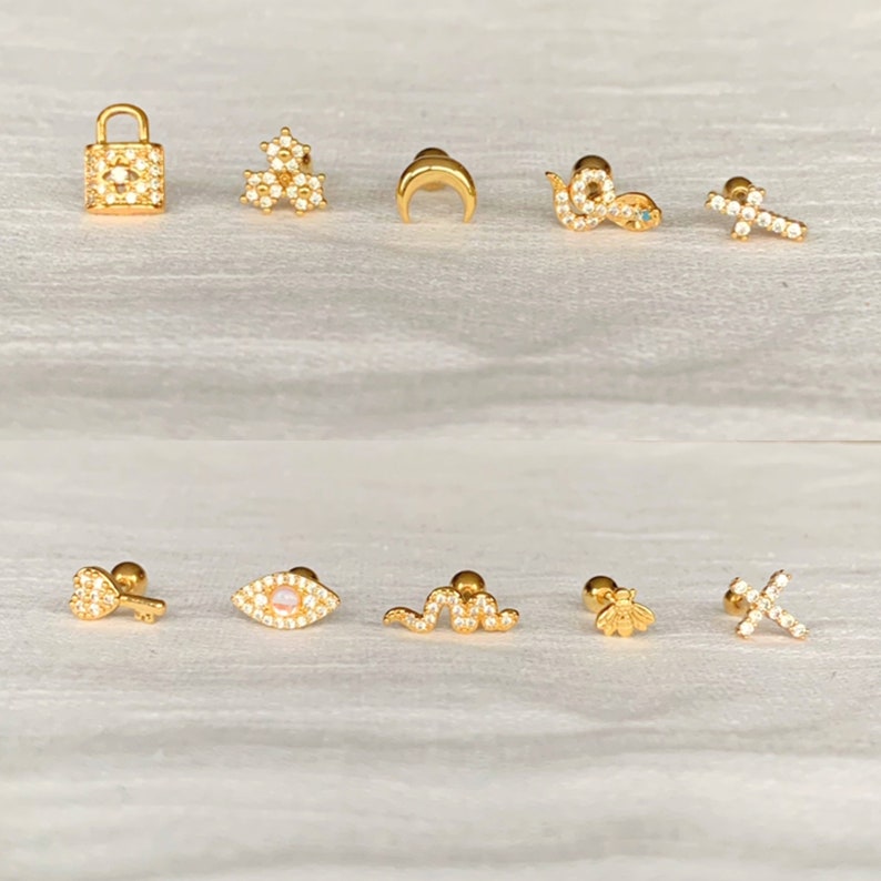 K&ouml;nnte beinhalten: Eine Sammlung goldfarbener Ohrringe mit verschiedenen Designs, darunter ein Schloss, Blumen, ein Halbmond, eine Schlange, ein Kreuz, ein Schl&uuml;ssel, ein Auge, eine Biene und mehr. Jeder Ohrring ist mit kleinen, klaren Edelsteinen besetzt.