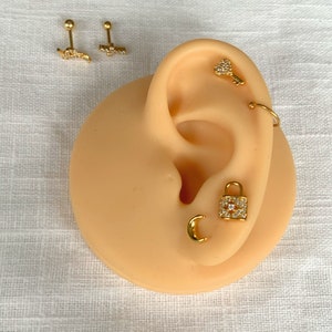 K&ouml;nnte beinhalten: Goldfarbene Ohrringe in verschiedenen Designs, darunter ein Schl&uuml;ssel, ein Schloss, ein Halbmond und ein gebogener Stab mit einer Reihe von Steinen. Die Ohrringe werden auf einem beigefarbenen Ohrmodell pr&auml;sentiert.