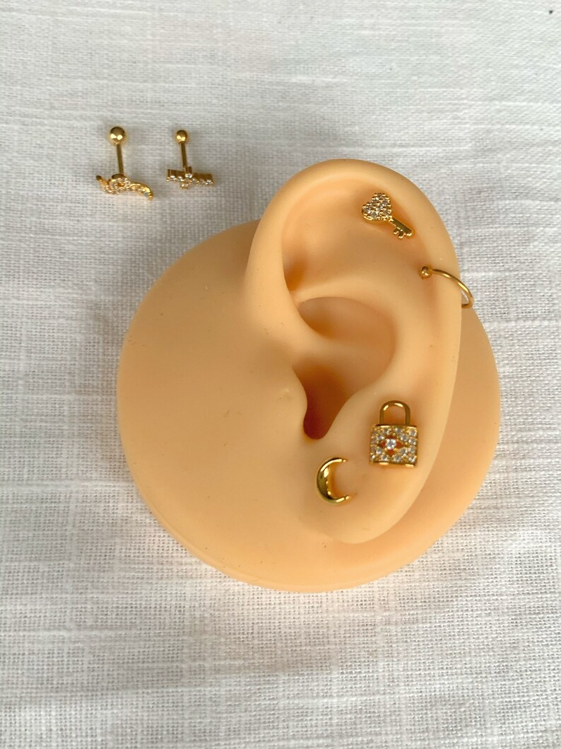 K&ouml;nnte beinhalten: Goldfarbene Ohrringe in verschiedenen Designs, darunter ein Schl&uuml;ssel, ein Schloss, ein Halbmond und ein gebogener Stab mit einer Reihe von Steinen. Die Ohrringe werden auf einem beigefarbenen Ohrmodell pr&auml;sentiert.