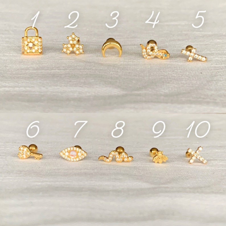 K&ouml;nnte beinhalten: Eine Sammlung von zehn goldfarbenen Ohrsteckern. Designs umfassen ein Vorh&auml;ngeschloss, Blumencluster, einen Halbmond, eine Schlange, ein Kreuz, einen Schl&uuml;ssel, ein Auge, eine Schlange, eine Biene und ein Kreuz. Jeder Ohrring ist mit kleinen, klaren Edelsteinen besetzt.