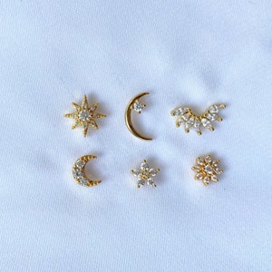 Könnte beinhalten: Eine Sammlung goldfarbener Schmuckstücke mit Himmelsmotiven, darunter Sterne, Monde und ein Sonnenstrahl-Design, alle mit klaren, funkelnden Edelsteinen verziert. Der Schmuck wird auf einer weißen Oberfläche präsentiert.