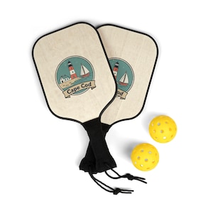 Puede incluir: Dos palas de pickleball con un diseño de Cape Cod y dos pelotas de pickleball amarillas. Las palas tienen un acabado de veta de madera clara con un mango negro y un gráfico de un faro y un velero. Las pelotas son amarillas con múltiples agujeros.