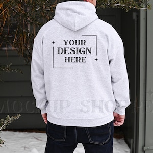 May include: A light gray hoodie with a hood and long sleeves. The back of the hoodie features the text "YOUR DESIGN HERE" inside a black rectangular frame. The person is wearing dark blue jeans.