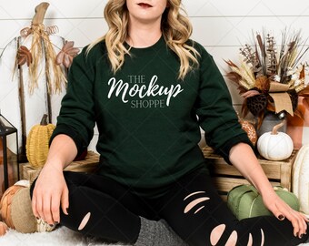 May include: A woman wearing a dark green sweatshirt with the text "The Mockup Shoppe" in white. She is sitting on a wooden bench with fall decorations, including pumpkins and gourds.
