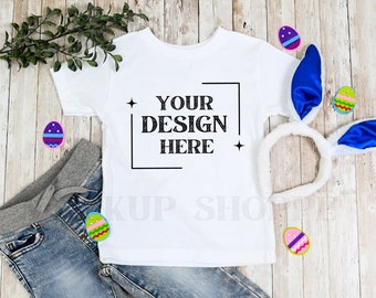 May include: White t-shirt with the text "YOUR DESIGN HERE" in a black box. The shirt is laying on a wooden surface with blue bunny ears, jeans, and colorful Easter eggs.