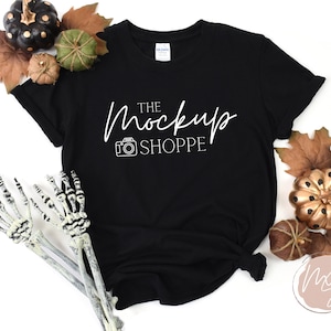 May include: Black t-shirt with white text that reads "The Mockup Shoppe" and a camera icon. The shirt is lying flat on a white surface with autumn-themed decorations.