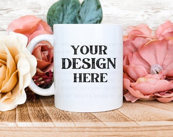 Könnte beinhalten: Weiße Keramiktasse mit einer schwarzen Aufschrift "YOUR DESIGN HERE" umgeben von künstlichen rosa und cremefarbenen Blumen auf einer Holzoberfläche.