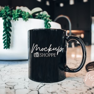 May include: A black ceramic mug with a white "Mockup Shoppe" logo and a camera icon. The mug is sitting on a white marble countertop.