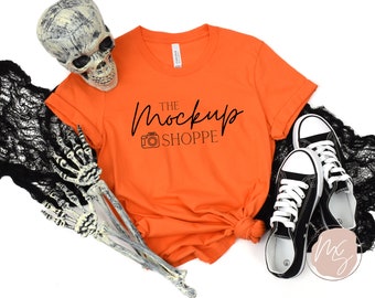 May include: Orange t-shirt with the text "THE MockUp SHOPPE" and a camera icon. The shirt is laid flat on a white background with black lace and a skeleton hand.