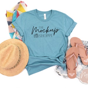 Op de afbeelding: Een turquoise t-shirt met de tekst "THE MockUp SHOPPE" en een camera-icoon bedrukt op de voorkant. Het shirt ligt op een witte achtergrond met een strohoed, een kleurrijke sjaal en een paar bruine slippers.
