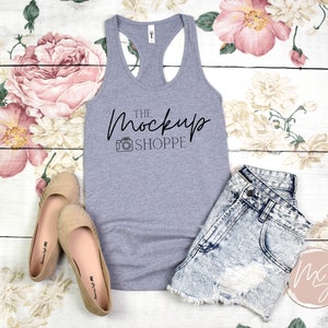 Op de afbeelding: Een grijs tanktop met een racerback-ontwerp. Het tanktop heeft de tekst "THE MOCKUP SHOPPE" in zwart op de voorkant gedrukt. Het tanktop ligt op een wit houten oppervlak met bloemenstof.