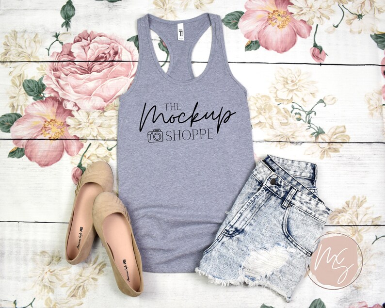 Op de afbeelding: Een grijs tanktop met een racerback-ontwerp. Het tanktop heeft de tekst "THE MOCKUP SHOPPE" in zwart op de voorkant gedrukt. Het tanktop ligt op een wit houten oppervlak met bloemenstof.