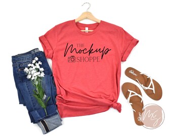 May include: A red t-shirt with the text "THE MOCKUP SHOPPE" and a camera icon, paired with blue jeans, white flowers, and brown sandals with white straps. The shirt is hanging on a wooden hanger.