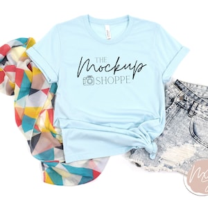 Könnte beinhalten: Ein hellblaues T-Shirt mit dem Text "The Mockup Shoppe" und einem Kamerasymbol auf der Vorderseite. Das T-Shirt liegt flach auf einem weißen Hintergrund mit einem bunten Schal und einer Jeans-Shorts.