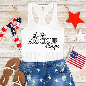 May include: White tank top with a black graphic that says "The Mock Up Shoppe" with a camera and heart design. The tank top is laying on a wooden surface with a red, white, and blue bandana, a pair of brown sandals, and a pair of blue denim shorts with white stars.