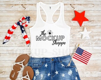 Op de afbeelding: Een wit tanktop met een zwarte afbeelding waarop "The Mock Up Shoppe" staat met een camera en een hart. Het tanktop ligt op een houten oppervlak met een rood-wit-blauwe bandana, een paar bruine sandalen en een blauwe spijkerbroek met witte sterren.
