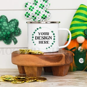May include: White enamel mug with a green shamrock design and the text "YOUR DESIGN HERE". The mug is sitting on a wooden coaster with gold coins scattered around it. A green and white shamrock and a green and white gnome are in the background.