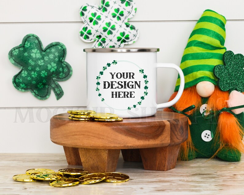 May include: White enamel mug with a green shamrock design and the text "YOUR DESIGN HERE". The mug is sitting on a wooden coaster with gold coins scattered around it. A green and white shamrock and a green and white gnome are in the background.