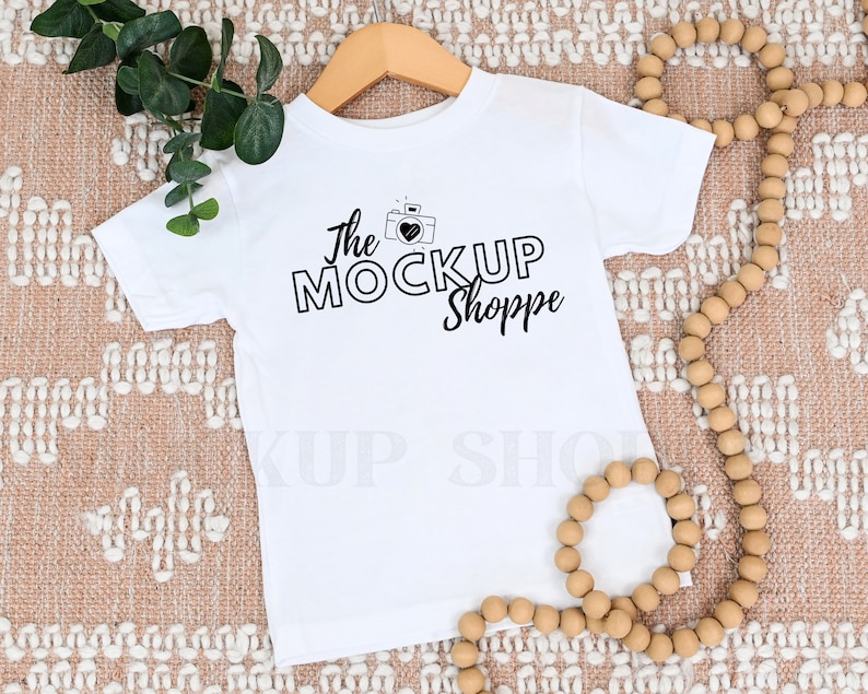 K&ouml;nnte beinhalten: Wei&szlig;es T-Shirt f&uuml;r Kleinkinder mit einem schwarzen Grafikdesign, auf dem "The Mock Up Shoppe" mit einem Kamerasymbol und einem Herzen steht.