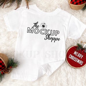 Puede incluir: Camiseta blanca con el texto "The Mockup Shoppe" y un gr&aacute;fico de c&aacute;mara. La camiseta est&aacute; sobre una superficie blanca y peluda con un adorno de cuadros rojos y negros y un tablero de letras rojo que dice "Feliz Navidad".