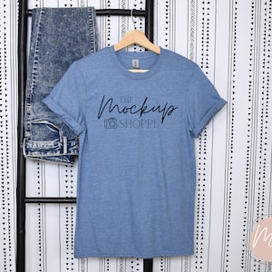 May include: A blue t-shirt with rolled up sleeves hanging on a wooden hanger. The t-shirt has the text "THE MockUp SHOPPE" printed on the front in black.
