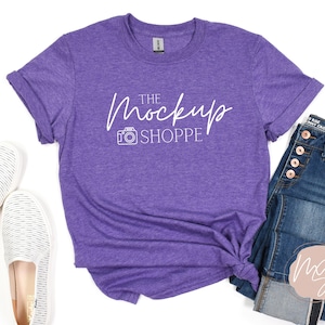 May include: A purple t-shirt with rolled-up sleeves and the text "THE MOCKUP SHOPPE" in white script. The shirt is styled with blue jeans and white slip-on shoes. The jeans have copper-colored buttons.