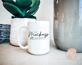 May include: White ceramic mug with a black handwritten logo that says "The Mockup Shoppe" with a camera icon. The mug is sitting on a light brown surface.