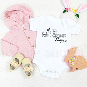 May include: A white baby onesie with short sleeves and a black graphic that says "The Mockup Shoppe" with a camera icon. The onesie is lying on a white fluffy blanket with a pink knitted cardigan, a pair of cream-coloured baby shoes, and a pink bunny headband with flowers.