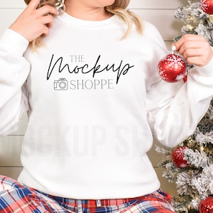 May include: White long-sleeve sweatshirt with the text "THE Mockup SHOPPE" and a camera icon. The model is wearing the sweatshirt with red and blue plaid pajama pants and is holding a red Christmas ornament.