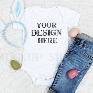 May include: A white baby onesie with the text "YOUR DESIGN HERE" on a white background. The onesie is lying on a white fluffy blanket with a blue and white bunny headband, a pair of blue jeans, and two Easter eggs.