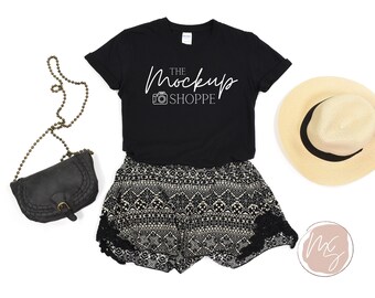 May include: Black t-shirt with the text "The Mockup Shoppe" and a camera icon. The shirt is paired with black and white patterned shorts, a black purse with a gold chain strap, and a straw hat.
