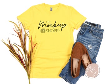 Op de afbeelding: Geel T-shirt met de tekst "THE MockUp SHOPPE" en een camera-icoon. Het shirt ligt op een wit oppervlak met een paar bruine loafers, een blauwe jeans en een tak herfstblad.