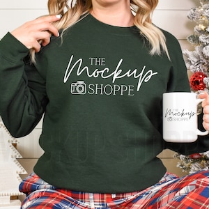 May include: A woman wearing a dark green sweatshirt with the text "THE MockUp SHOPPE" and a white camera icon. She is holding a white mug with the same text and icon. The woman is wearing red and black plaid trousers and is sitting in front of a white wooden wall with a decorated Christmas tree behind her.