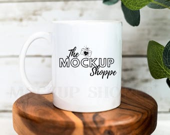 May include: White ceramic mug with a black design that says "The MockUp Shoppe" with a camera icon and a heart. The mug is sitting on a wooden surface.