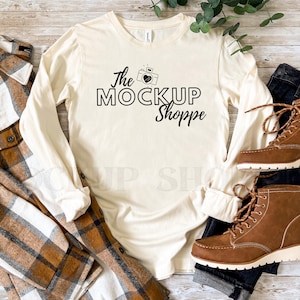 May include: A beige long-sleeve t-shirt with the text "The MockUp Shoppe" and a camera graphic. The shirt is laid out on a wooden surface with a plaid shirt, jeans, and brown boots.
