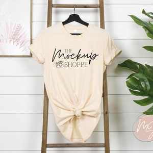 May include: A beige t-shirt mockup with the text "THE MockUp SHOPPE" and a camera icon printed on the front. The shirt is hanging on a wooden ladder against a white wood panel background.