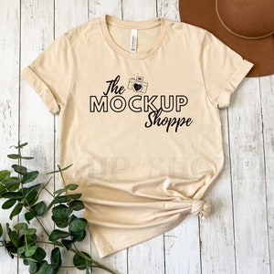 May include: A beige t-shirt with a black graphic design that says "The Mock Up Shoppe" with a camera and heart icon. The t-shirt is laid flat on a white wooden surface.