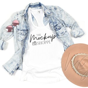 May include: White tank top with a black graphic that says "THE MOCKUP SHOPPE" with a camera icon. The tank top is laying on a white surface with a light blue denim jacket and a brown hat with a pearl trim.