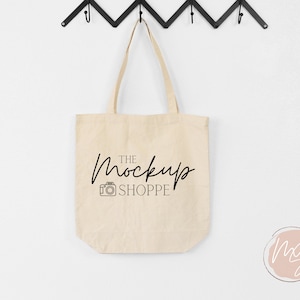 May include: A beige canvas tote bag with black handles hanging on hooks. The bag has a black camera icon and the text "THE Mockup SHOPPE" printed on the front.