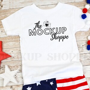 May include: White t-shirt with a black graphic of a camera with a heart inside and the text "The Mockup Shoppe". The shirt is laying on a wooden surface with red, white, and blue decorations.