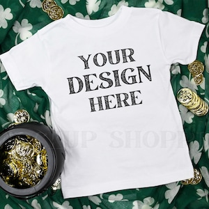 May include: White t-shirt with the text "YOUR DESIGN HERE" in black. The shirt is on a green fabric with white shamrocks, next to a black pot filled with gold coins and scattered gold coins.